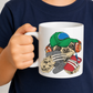 A child in a navy blue shirt holding a white ceramic mug. The mug features a cartoon sloth graphic wearing a green cap, orange headphones, and holding a gray game controller and a red phone. Purchase your mug from Coffee and Moo NZ.