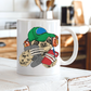 A white ceramic mug with a colorful cartoon sloth graphic wearing a green cap, orange headphones, and holding a gaming controller and a red phone, sitting on a rustic white wooden table with a blurred kitchen background. Purchase your mug from Coffee and Moo NZ.