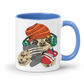 A white ceramic mug with a blue interior and handle, featuring a cartoon sloth wearing an orange knit beanie, green headphones, and holding a red phone and gray game controller. Grab your gaming sloth mug from Coffee and Moo NZ.