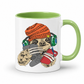 A white ceramic mug with a light green interior and handle, featuring a cartoon graphic of a sloth wearing an orange knit beanie, green headphones, and holding a grey game controller and a red phone. Grab your gaming sloth mug from Coffee and Moo NZ.