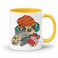 A white ceramic coffee mug with a yellow interior and handle, featuring a cartoon sloth wearing an orange knit beanie, green headphones, and holding a grey game controller and a red phone. Grab your gaming sloth mug from Coffee and Moo NZ.