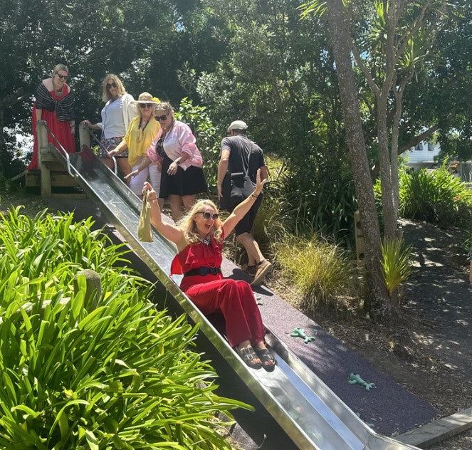 A woman in a red dress slides down a metal slide in an outdoor park setting, surrounded by lush green plants and trees, with other people standing on a wooden platform behind her. Pick a card from the 'Getting Lost Girls Road Trip Edition' game. Pack Includes 26 direction and activity cards, ready for a weekend getaway. Purchase yours from Coffee and Moo NZ