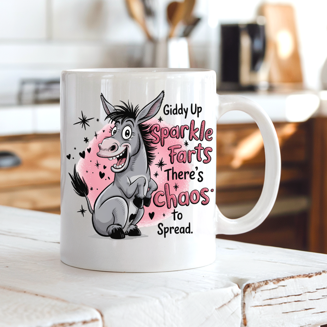 A white ceramic coffee mug with a cartoon donkey and the text 'Giddy Up Sparkle Farts There's Chaos to Spread.' sitting on a rustic white wooden table with a blurred kitchen background. from Coffee and Moo.