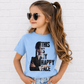 A young girl with long brown hair in a ponytail, wearing heart-shaped sunglasses and a light blue graphic t-shirt with a black and white print of Wednesday Addams and the text 'THIS IS MY HAPPY FACE'. She is posing in front of a white brick wall. Grab your kids tee from Coffee and Moo NZ.