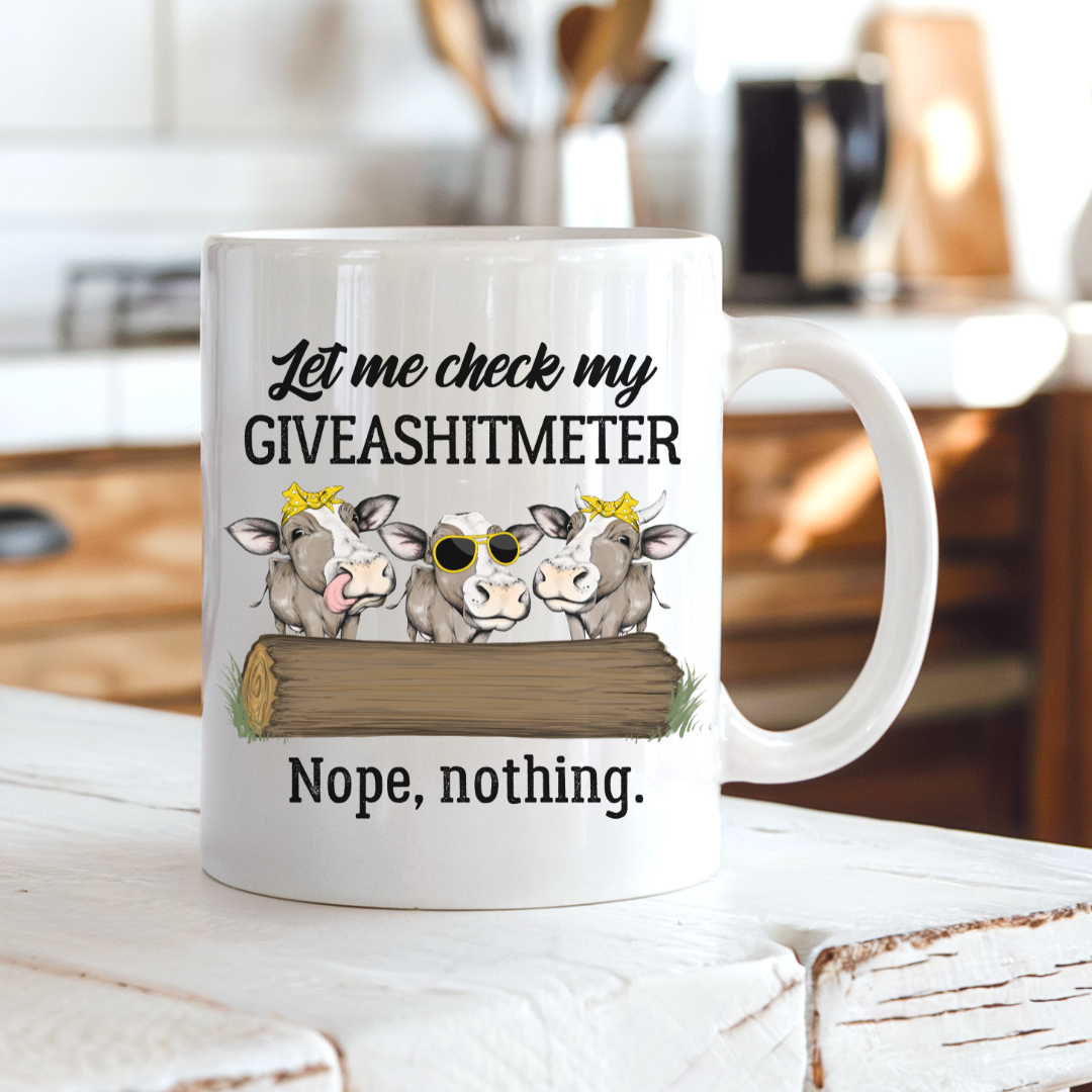 A white ceramic coffee mug with a humorous graphic and text, displayed on a rustic white wooden table with a blurred kitchen background. Grab your cheeky mug from Coffee and Moo NZ.