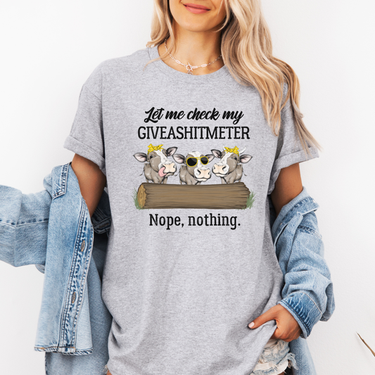 A woman wearing a grey graphic t-shirt with a cow illustration and the text 'Let me check my GIVEASHITMETER Nope, nothing.' She has a light blue denim jacket draped off her shoulders against a plain white background. Grab your from Coffee and Moo NZ.