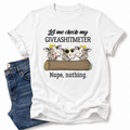 A white graphic t-shirt featuring a cartoon design of three cows and the text 'Let me check my GIVEASHITMETER Nope, nothing.' styled flat-lay next to a pair of blue jeans on a white background. Grab your from Coffee and Moo NZ.