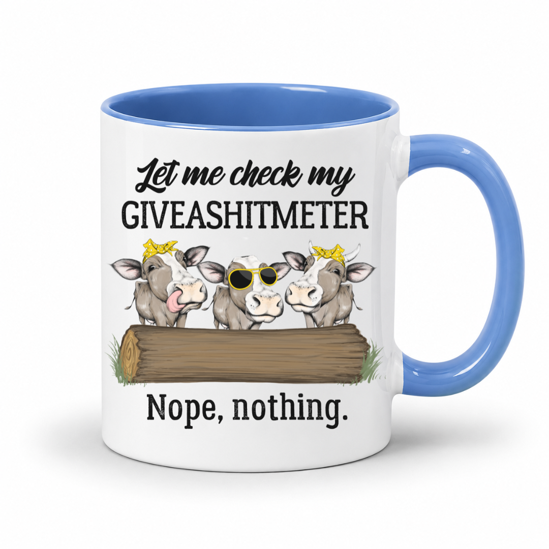 A white ceramic mug with a blue handle and interior, featuring a humorous graphic of three cows behind a log with the text 'Let me check my GIVEASHITMETER Nope, nothing.'. Grab your cheeky mug from Coffee and Moo NZ.