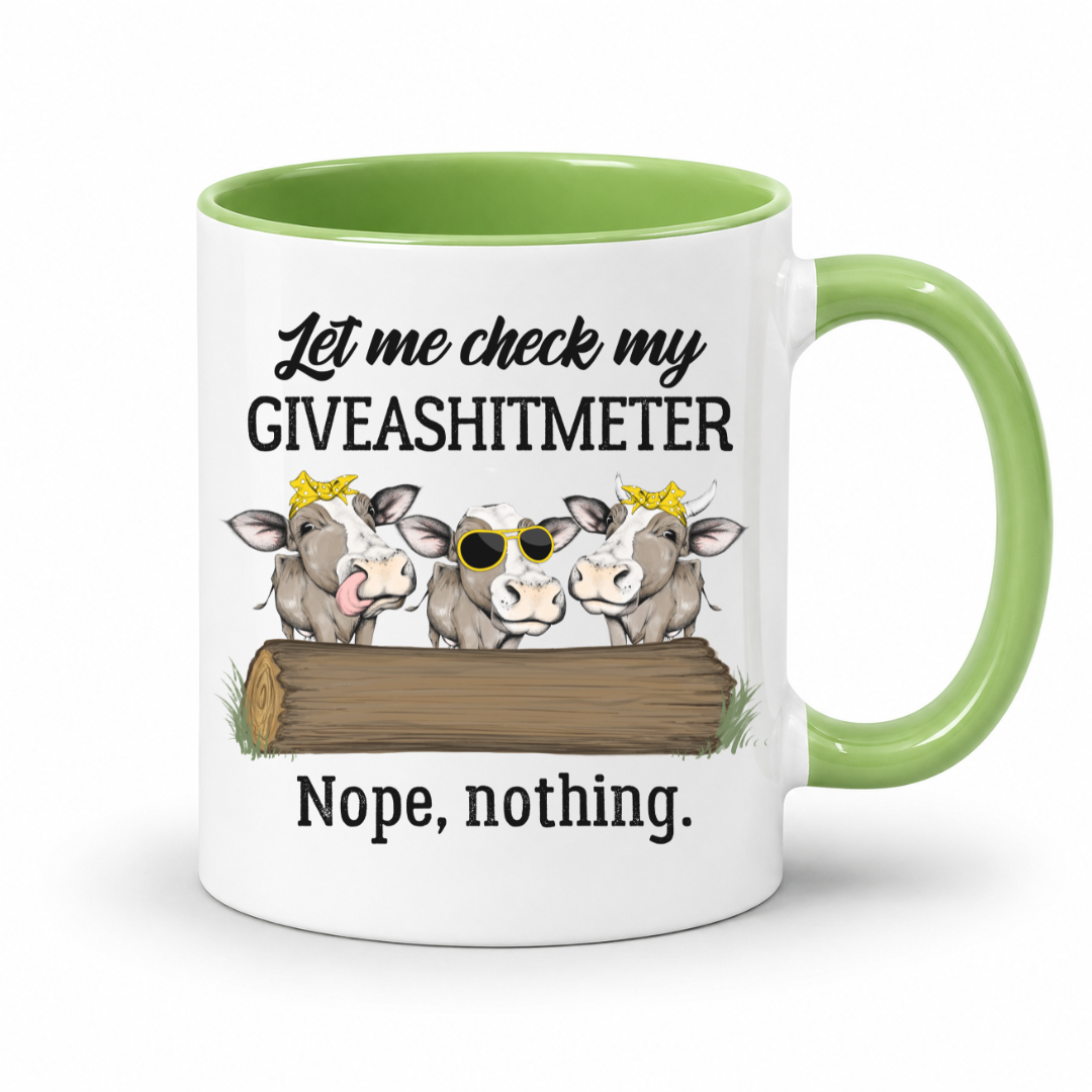 A white ceramic mug with a light green interior and handle, featuring a humorous graphic of three cartoon cows behind a log with the text 'Let me check my GIVEASHITMETER Nope, nothing.'. Grab your cheeky mug from Coffee and Moo NZ.