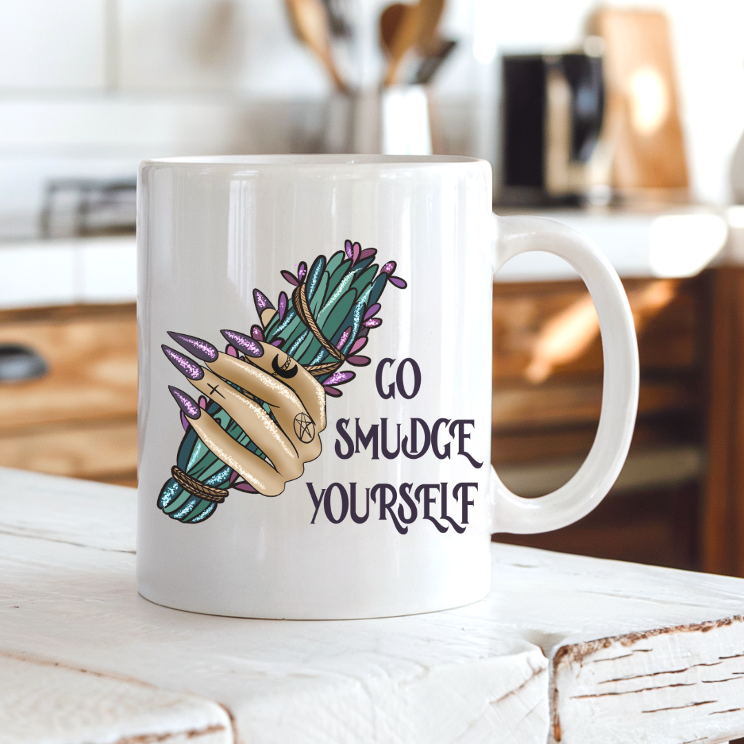 A white ceramic mug with a hand graphic holding a green smudge bundle and the text 'GO SMUDGE YOURSELF' in black, set on a rustic white wooden table with a blurred kitchen background. Purchase from Coffee and Moo NZ.