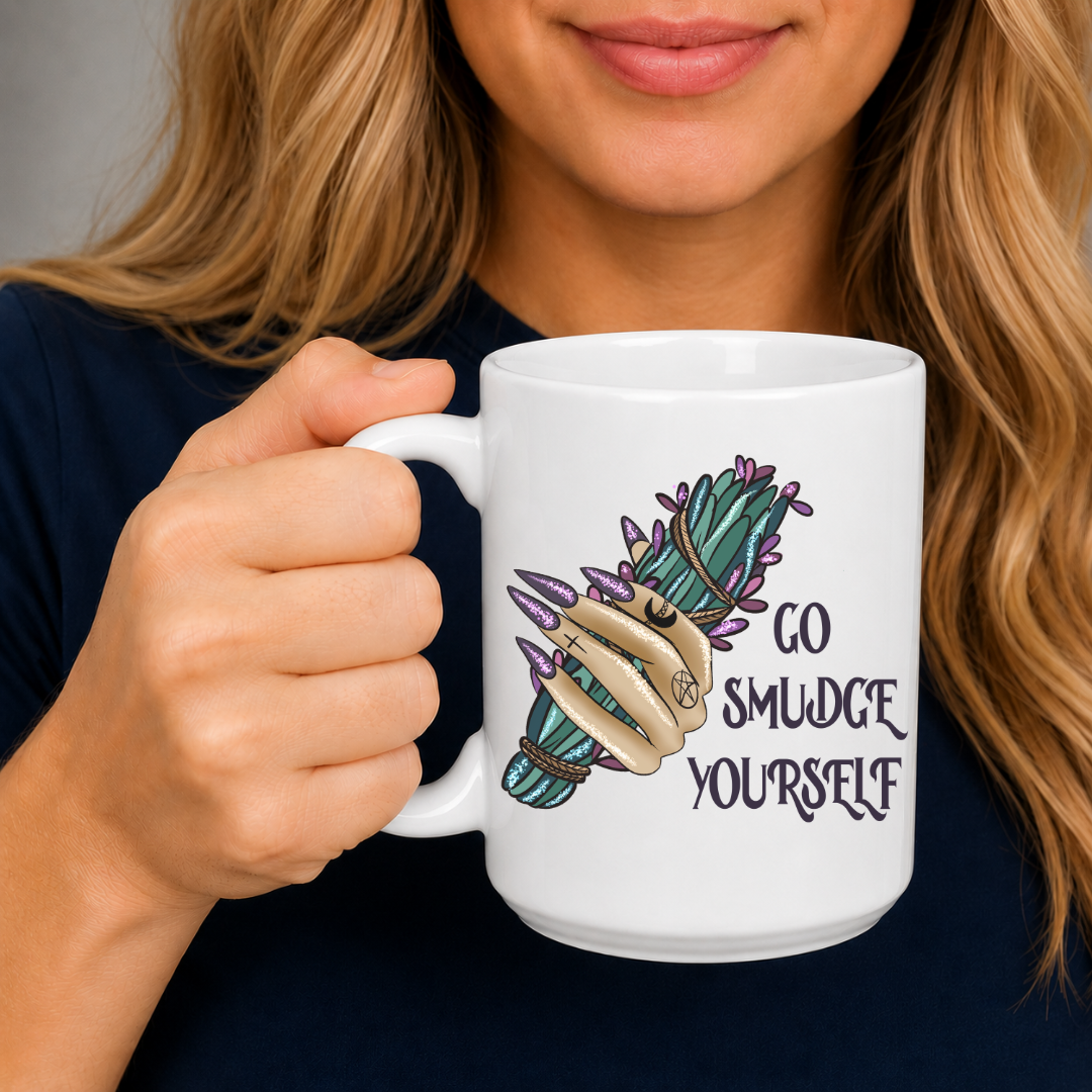 A woman holding a white ceramic mug with a graphic of a hand holding a green and purple smudge bundle next to the text 'GO SMUDGE YOURSELF'. Purchase from Coffee and Moo NZ.