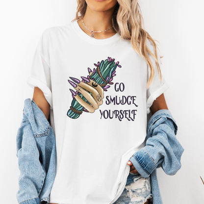 A woman wearing a white graphic t-shirt with a hand holding a green and purple smudge bundle and the text 'GO SMUDGE YOURSELF' printed on the front. She has a light blue denim jacket draped off her shoulders and is posing against a plain white background. Purchase from Coffee and Moo NZ.
