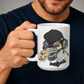 A man holding a white ceramic mug featuring a cartoon illustration of a sloth wearing a black knit beanie, over-ear headphones, and holding a cup with the word 'TEARS' on it and a diary,  Grab yours from Coffee and Moo NZ.