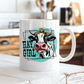A white ceramic coffee mug with a cartoon cow graphic and the text 'HAY GIRL' on the front, set on a rustic white wooden table with a blurred kitchen background.  from Coffee and Moo NZ.