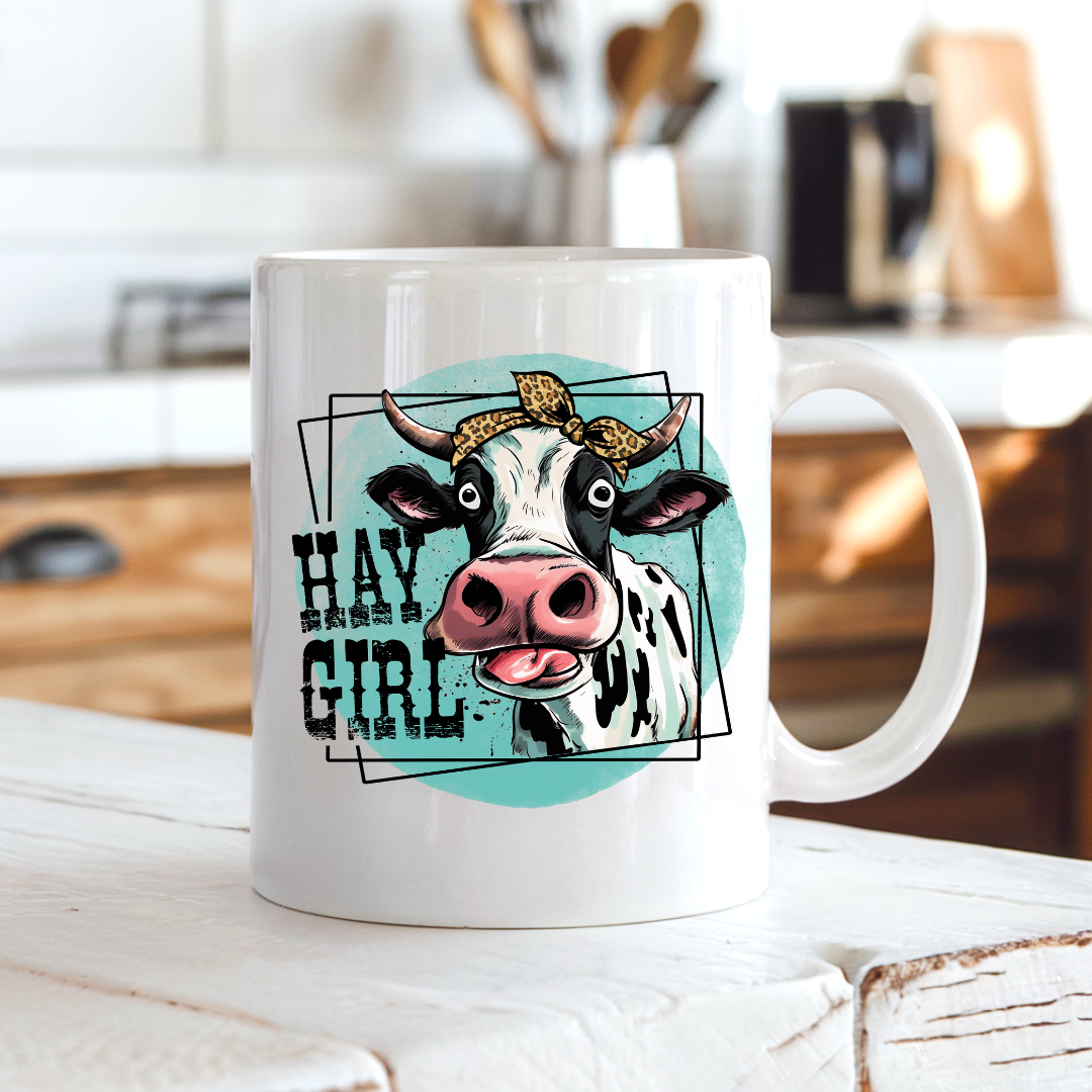 A white ceramic coffee mug with a cartoon cow graphic and the text 'HAY GIRL' on the front, set on a rustic white wooden table with a blurred kitchen background.  from Coffee and Moo NZ.