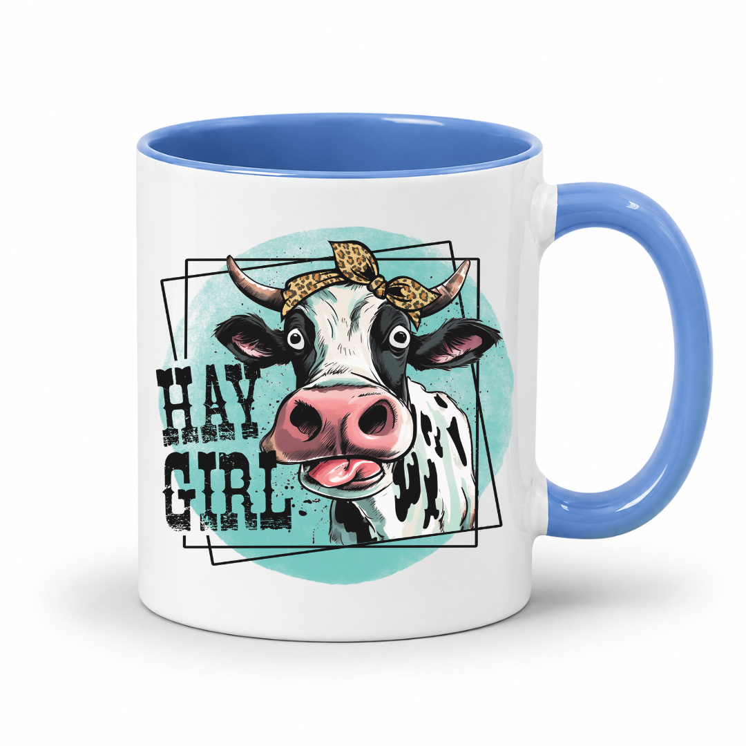A white ceramic coffee mug with a blue handle and interior, featuring a graphic illustration of a black-and-white cow with a leopard-print bow and the text 'HAY GIRL' on a light blue circular background.  from Coffee and Moo NZ.