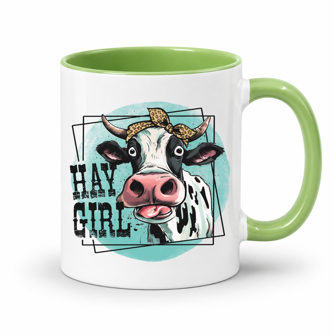 A white ceramic coffee mug with a light green interior and handle, featuring a graphic illustration of a cow wearing a leopard-print bow and the text 'HAY GIRL' on a light blue circular background.  from Coffee and Moo NZ.