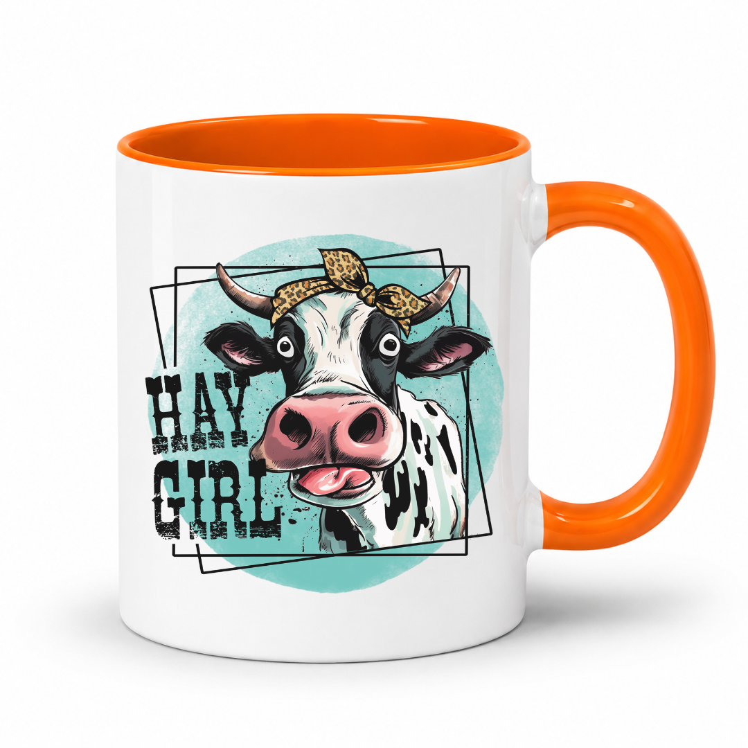 A white ceramic coffee mug with a bright orange handle and interior, featuring a graphic illustration of a cow wearing a leopard-print headband on a light blue circular background, with the text 'HAY GIRL' in black.  from Coffee and Moo NZ.