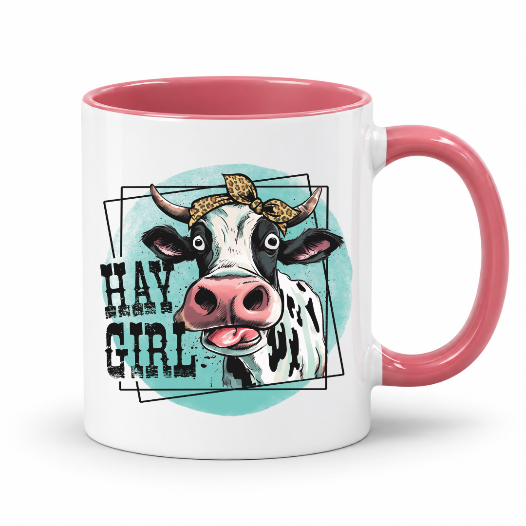 A white ceramic mug with a pink interior and handle, featuring a graphic of a black-and-white cow wearing a leopard-print bandana and the text 'HAY GIRL' on a light blue oval background.  from Coffee and Moo NZ.