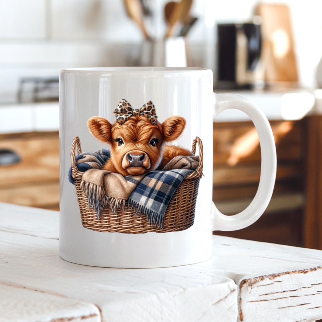 A white ceramic mug with a printed illustration of a cute brown calf in a wicker basket, set on a rustic white wooden table with a blurred kitchen background. from Coffee and Moo.