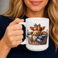 A person holding a white ceramic mug with a printed illustration of a brown cow calf in a basket with a plaid blanket and a leopard-print bow. The person is wearing a dark navy shirt. from Coffee and Moo.