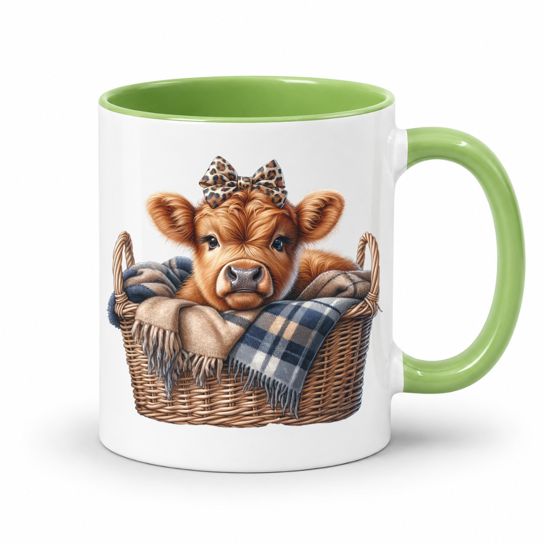 A white ceramic mug with a green interior and handle, featuring a printed illustration of a cute brown calf sitting in a wicker basket with a plaid blanket and leopard-print bow. from Coffee and Moo NZ.