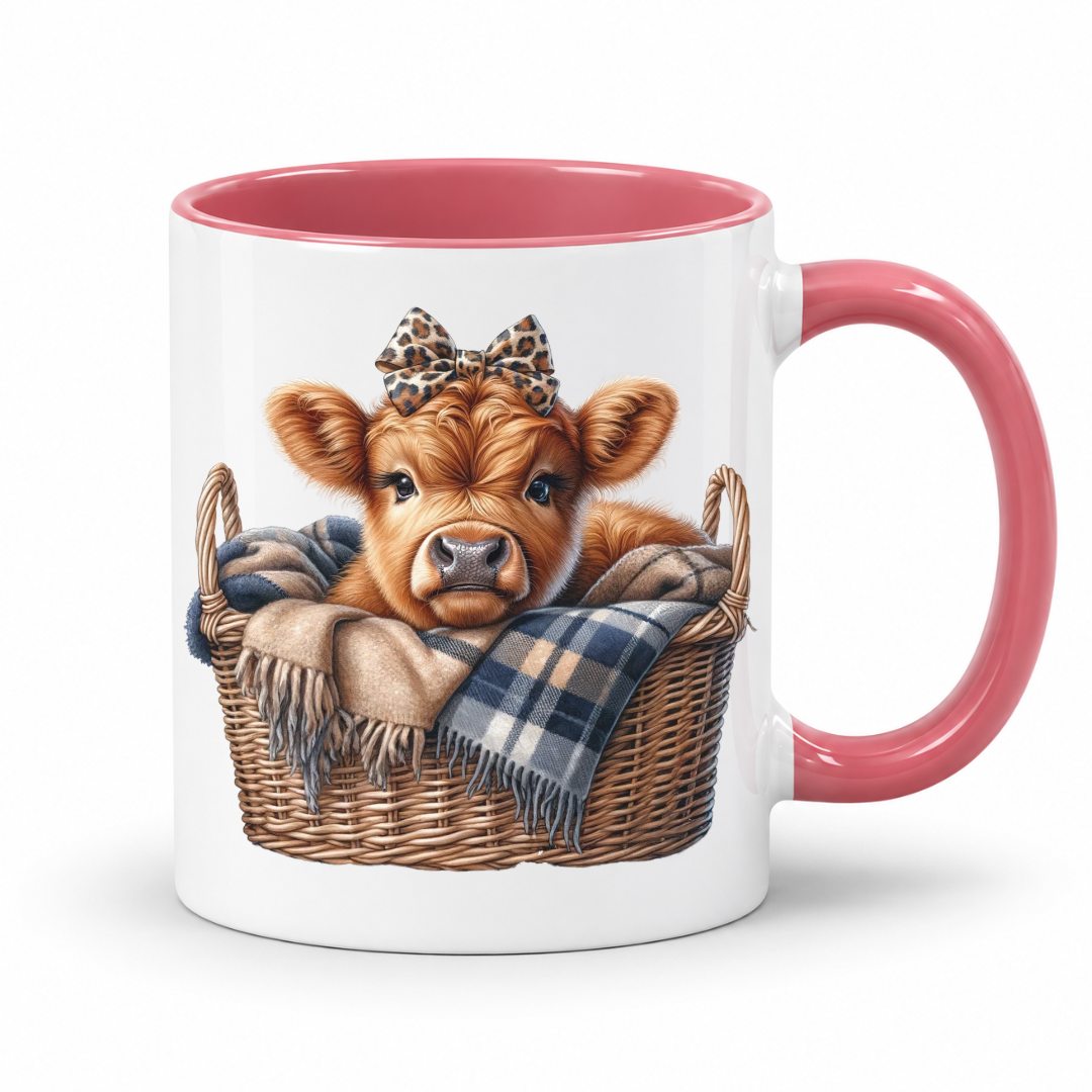 A white ceramic mug with a pink interior and handle, featuring a printed illustration of a brown calf in a wicker basket with a plaid blanket and leopard-print bow. from Coffee and Moo NZ.