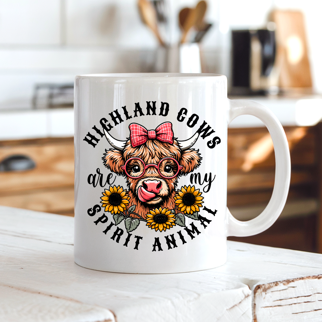 A white ceramic mug with a cartoon Highland cow graphic and the text 'HIGHLAND COWS are my SPIRIT ANIMAL', shown on a white wooden surface with a blurred kitchen background. from Coffee and Moo NZ.
