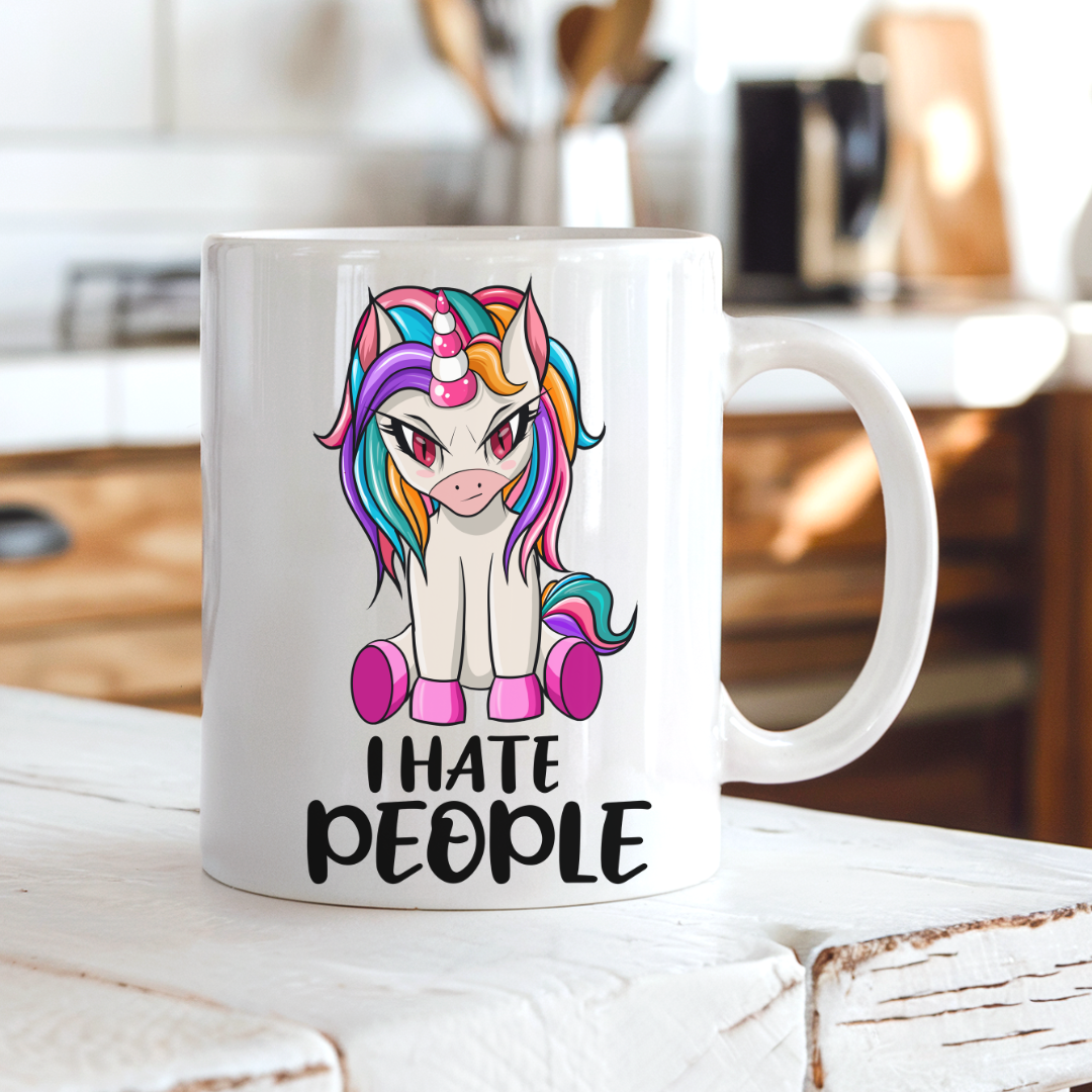 A white ceramic mug with a colorful cartoon unicorn graphic and the text 'I HATE PEOPLE' printed on the front, set on a rustic white wooden table with a blurred kitchen background. From Coffee and Moo NZ.