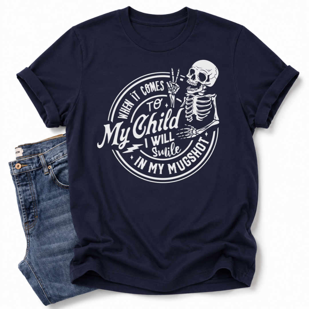 A navy blue short-sleeve t-shirt with a white graphic print of a skeleton and the text 'When it comes to My Child I Will Smile in my mugshot', styled flat-lay next to a pair of blue jeans on a white background. Grab your tee from Coffee and Moo NZ.