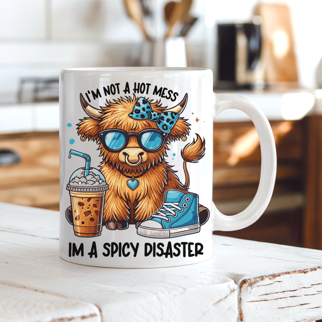 A white ceramic coffee mug with a colorful cartoon illustration of a fluffy brown highland cow wearing blue sunglasses and a blue bow tie, next to an iced coffee and a blue sneaker. The text reads 'I'M NOT A HOT MESS' above the cow and 'I'M A SPICY DISASTER' below. from Coffee and Moo NZ.