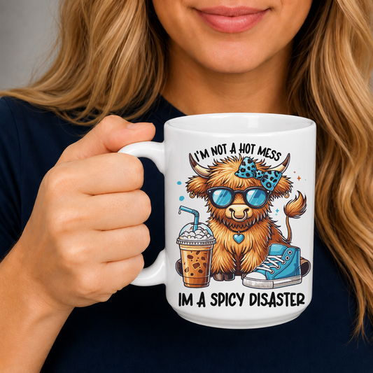 A person holding a white ceramic mug with a colorful cartoon illustration of a fluffy brown creature wearing blue sunglasses and a blue patterned headband, with the text 'I'M NOT A HOT MESS' above and 'I'M A SPICY DISASTER' below. from Coffee and Moo NZ.