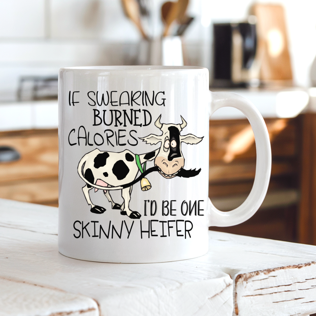 A white ceramic coffee mug with a cartoon cow and the text 'IF SWEARING BURNED CALORIES I'D BE ONE SKINNY HEIFER' placed on a rustic white wooden table with a blurred kitchen background. from Coffee and Moo NZ.