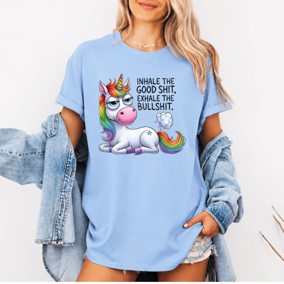 A woman wearing a light blue graphic t-shirt with a cartoon unicorn and the text 'INHALE THE GOOD SHIT, EXHALE THE BULLSHIT.' She has a light-wash denim jacket off her shoulders against a plain white background.  Purchase from Coffee and Moo NZ.