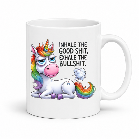 A white ceramic mug featuring a cartoon illustration of a white unicorn with a rainbow mane, horn, and tail, sitting next to the black text 'INHALE THE GOOD SHIT, EXHALE THE BULLSHIT.' on a white background. Purchase from Coffee and Moo NZ.