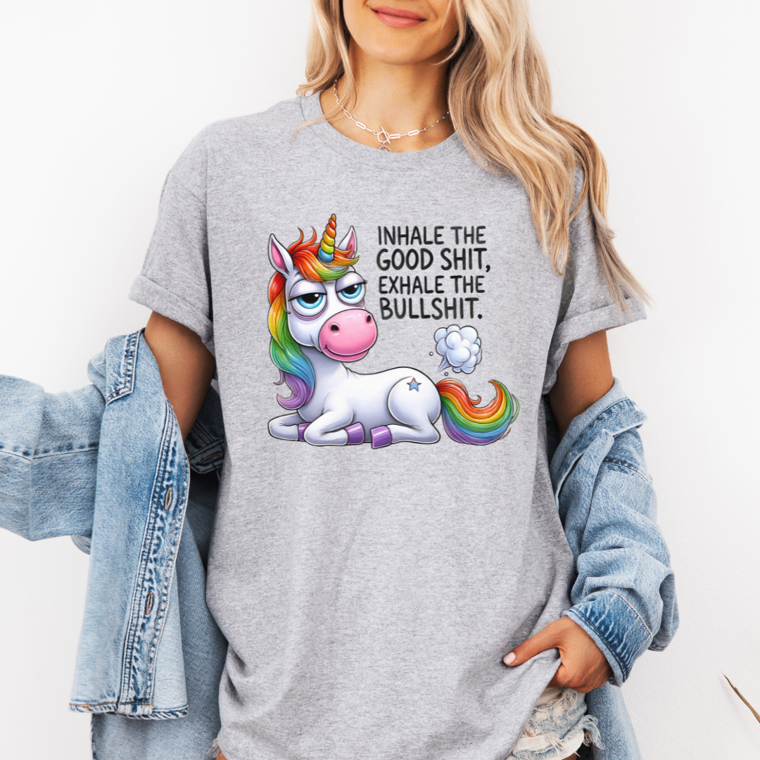 A woman wearing a grey graphic t-shirt with a cartoon rainbow unicorn and the text 'INHALE THE GOOD SHIT, EXHALE THE BULLSHIT.' She has a light blue denim jacket draped off her shoulders against a plain white background. Purchase from Coffee and Moo NZ.