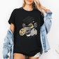 A woman wearing a black short-sleeve graphic t-shirt with a stylized Emo sloth wearing a beanie, headphones, and holding a coffee cup with the word "TEARS" on it. Grab your tee from Coffee and Moo NZ.