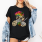 A woman wearing a black unhinged adult graphic tee features our resident zen master—a delightfully baked sloth rocking a vibrant, colourful tie-dye beanie topped with a cannabis leaf, proudly clutching a bong and a jar of premium green buds. Grab your sloth stoner tee from Coffee and Moo NZ.
