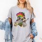 A woman wearing a grey unhinged adult graphic tee features our resident zen master—a delightfully baked sloth rocking a vibrant, colourful tie-dye beanie topped with a cannabis leaf, proudly clutching a bong and a jar of premium green buds. Grab your sloth stoner tee from Coffee and Moo NZ.
