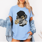 A woman wearing an oversized light blue graphic t-shirt with a cartoon slot wearing a black beanie and headphones. She has a light-wash denim jacket off her shoulders and is posing against a clean white background. Grab your emo sloth tee from Coffee and Moo NZ.