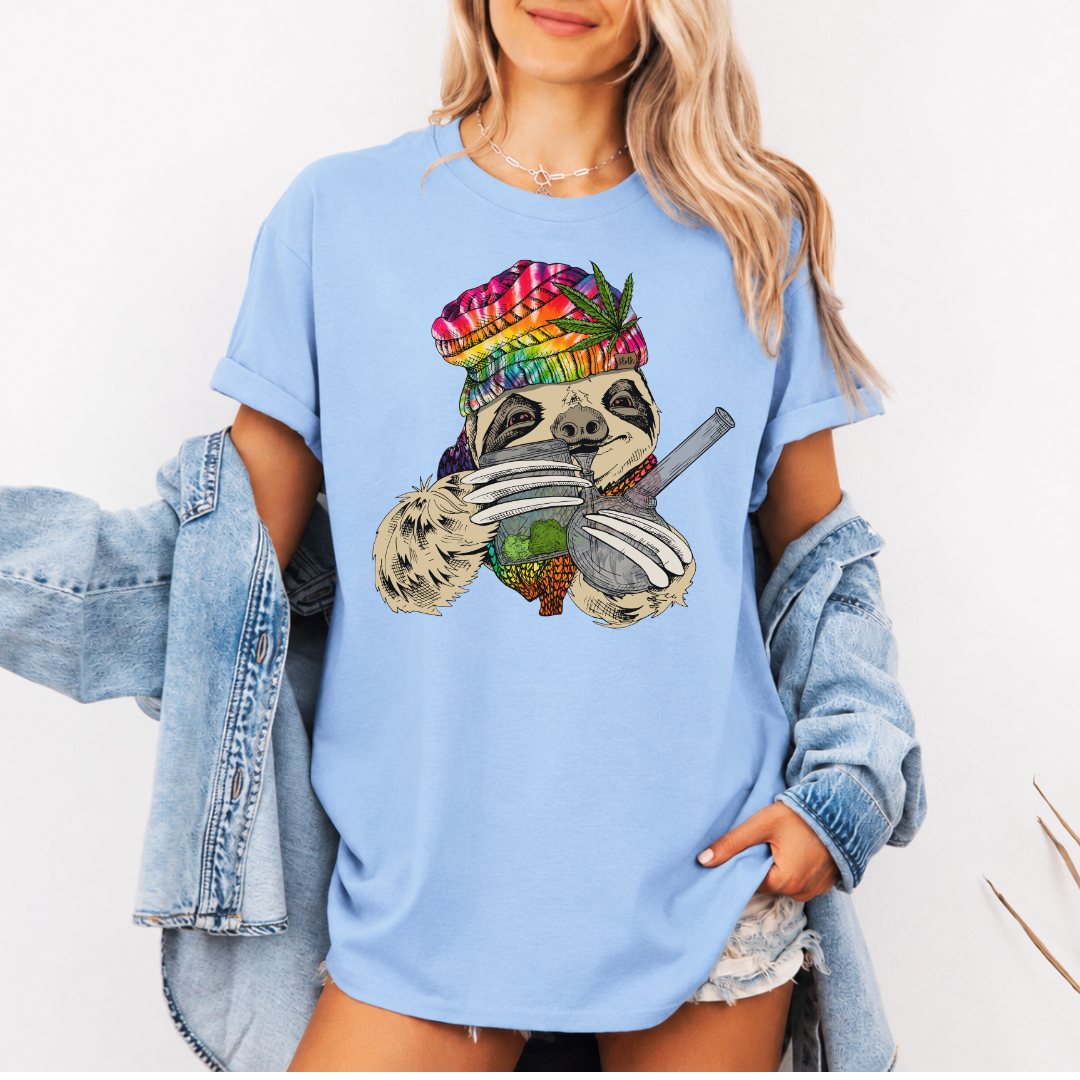 A woman wearing a baby blue unhinged adult graphic tee features our resident zen master—a delightfully baked sloth rocking a vibrant, colourful tie-dye beanie topped with a cannabis leaf, proudly clutching a bong and a jar of premium green buds. Grab your sloth stoner tee from Coffee and Moo NZ.