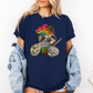 A woman wearing a navy unhinged adult graphic tee features our resident zen master—a delightfully baked sloth rocking a vibrant, colourful tie-dye beanie topped with a cannabis leaf, proudly clutching a bong and a jar of premium green buds. Grab your sloth stoner tee from Coffee and Moo NZ.