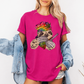 A woman wearing a pink unhinged adult graphic tee features our resident zen master—a delightfully baked sloth rocking a vibrant, colourful tie-dye beanie topped with a cannabis leaf, proudly clutching a bong and a jar of premium green buds. Grab your sloth stoner tee from Coffee and Moo NZ.