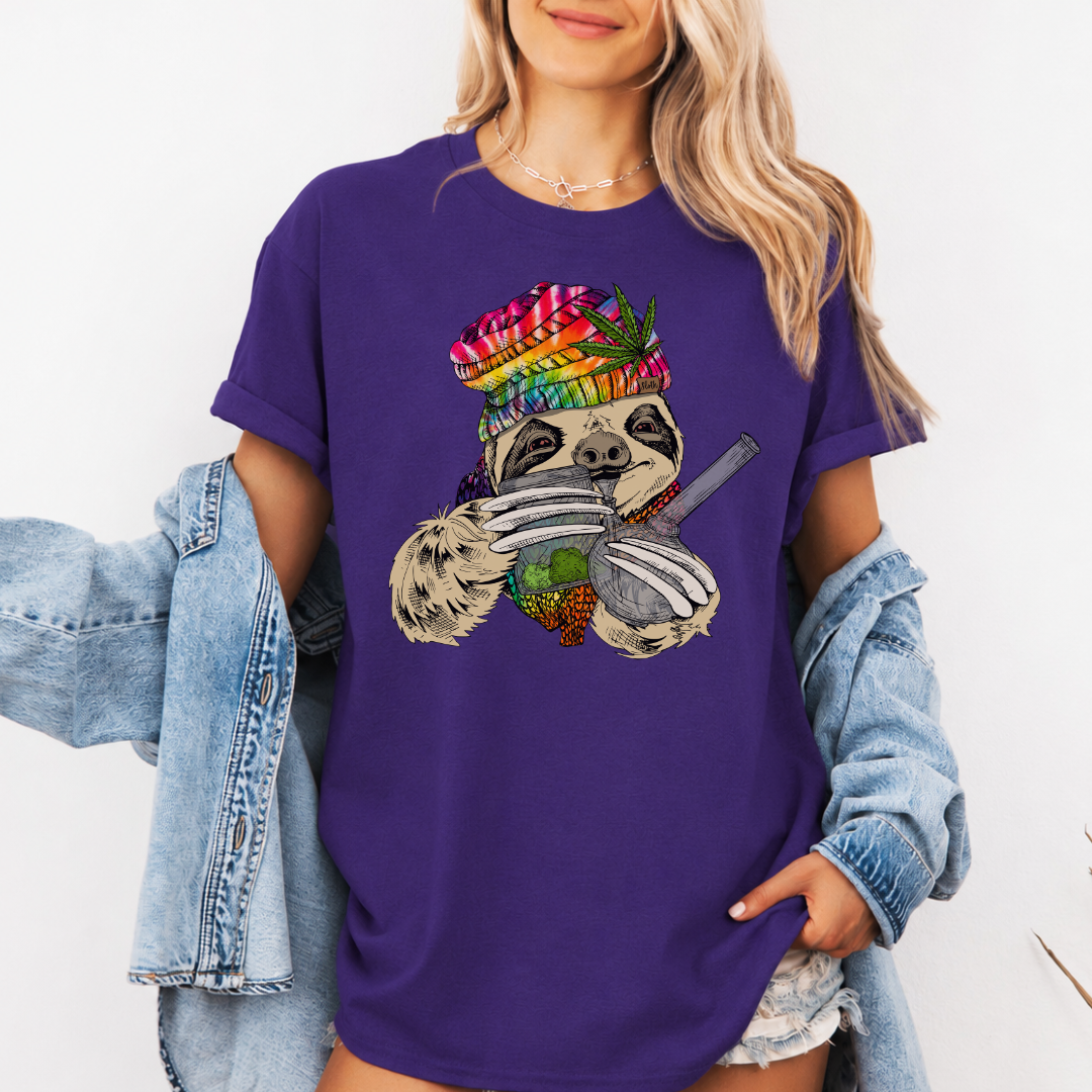 A woman wearing a purple unhinged adult graphic tee features our resident zen master—a delightfully baked sloth rocking a vibrant, colourful tie-dye beanie topped with a cannabis leaf, proudly clutching a bong and a jar of premium green buds. Grab your sloth stoner tee from Coffee and Moo NZ.