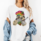 A woman wearing a white unhinged adult graphic tee features our resident zen master—a delightfully baked sloth rocking a vibrant, colourful tie-dye beanie topped with a cannabis leaf, proudly clutching a bong and a jar of premium green buds. Grab your sloth stoner tee from Coffee and Moo NZ.