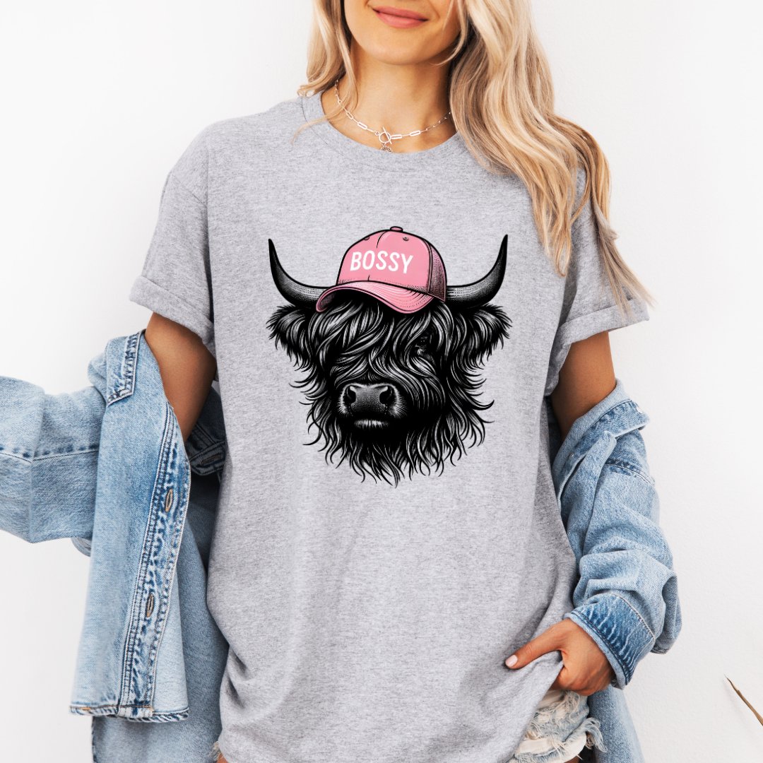A woman wearing a grey oversized t-shirt with a graphic of a black fluffy Highland cow wearing a pink cap that says 'BOSSY' in white text. She has a light blue denim jacket draped off her shoulders against a plain white background. Grab your tee from Coffee and Moo NZ.