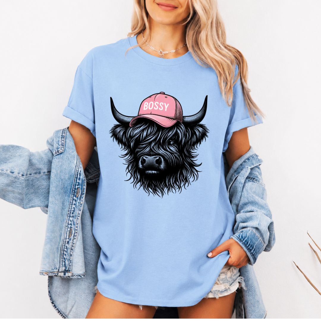A woman wearing an oversized light blue t-shirt with a graphic of a black highland cow wearing a pink cap that says 'BOSSY'. She has a light-wash denim jacket off her shoulders and is posing against a white background.  Grab your tee from Coffee and Moo NZ.