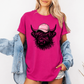 A woman wearing a bright pink graphic t-shirt featuring a black and pink highland cow wearing a pink cap with the word 'BOSSY' in white. She has a light blue denim jacket draped off her shoulders and is posing against a clean white background.  Grab your tee from Coffee and Moo NZ.