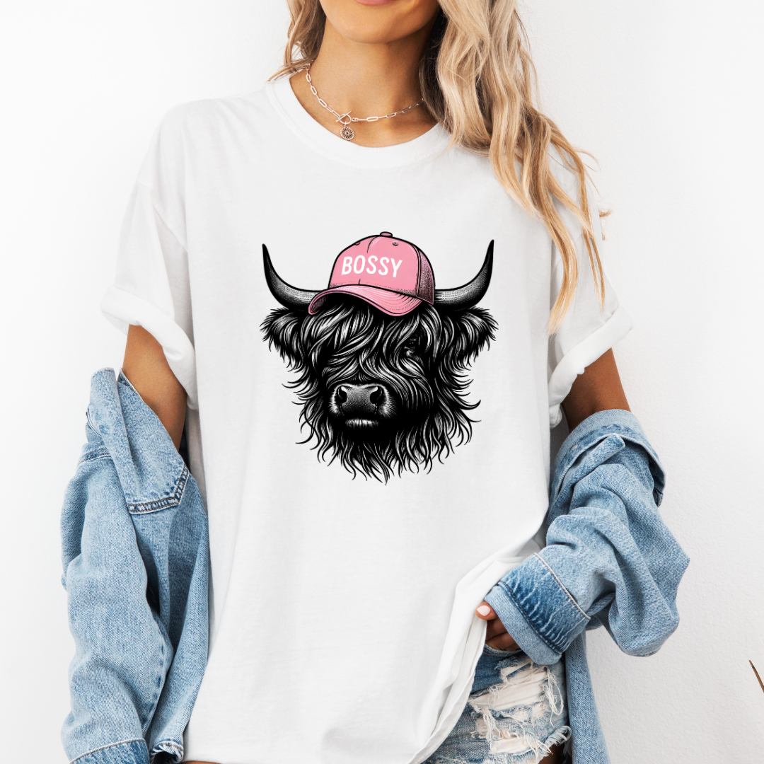 A woman wearing a white graphic t-shirt with a black highland cow illustration wearing a pink 'BOSSY' cap. She has a light blue denim jacket draped off her shoulders and is posing against a clean white background.  Grab your tee from Coffee and Moo NZ.