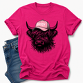 A bright pink short-sleeve t-shirt with a graphic of a shaggy Highland cow wearing a pink cap that says 'BOSSY' in white letters. The t-shirt is styled flat-lay with a pair of blue jeans on a clean white background.  Grab your tee from Coffee and Moo NZ.